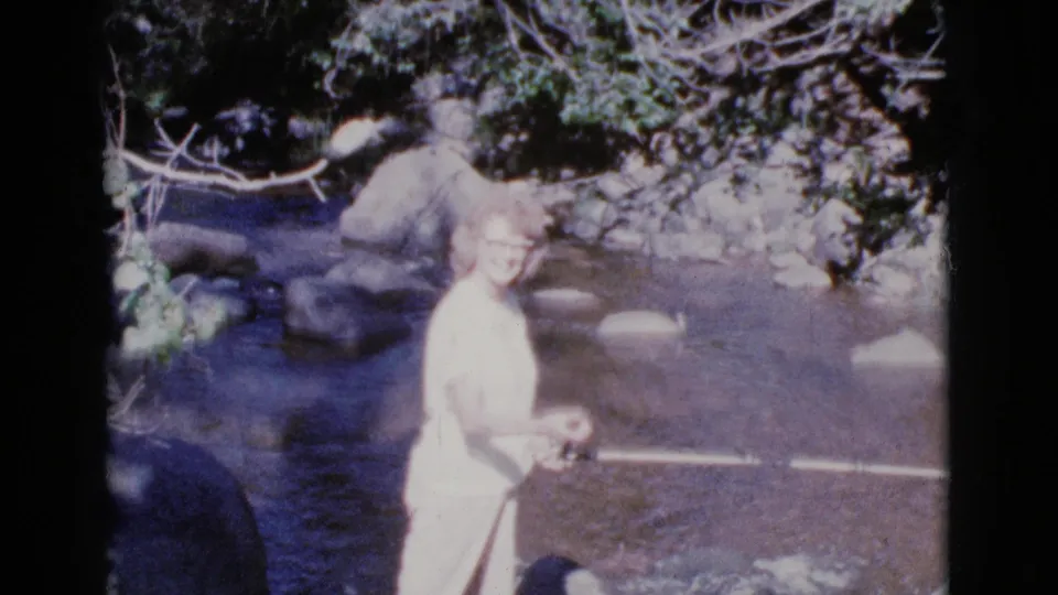 A woman in a white dress turns toward the camera with a fishing rod lowered across the shallow river in 1964 Cottonwood, Arizona footage.