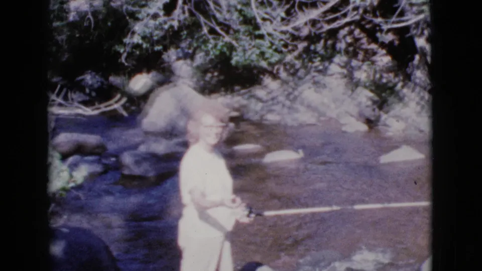 The woman stands in the shallow Cottonwood river setting with the rod extended, surrounded by rocks, water, and dense bank vegetation.