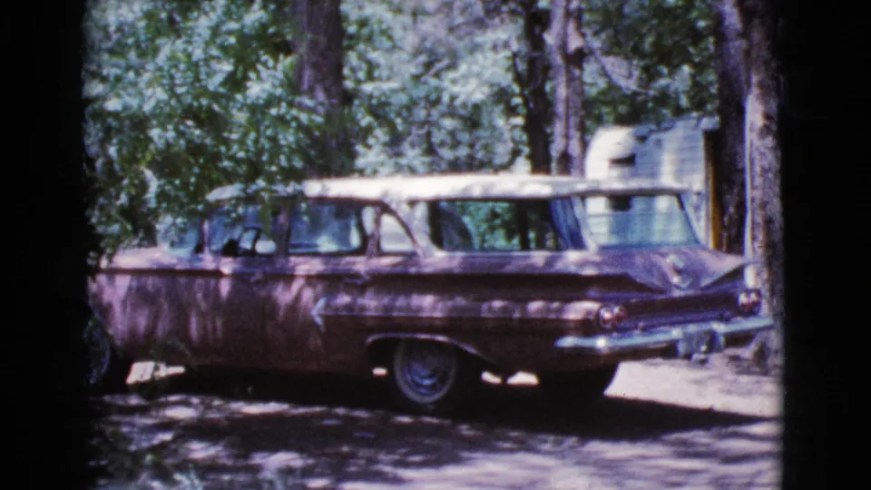 A mid-century station wagon is parked in a wooded campsite area, with a camper visible behind the car.