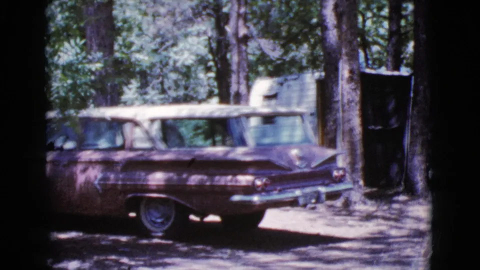 The station wagon remains parked beneath tall trees, with the camper and campsite surroundings visible in 1964 home-movie footage.