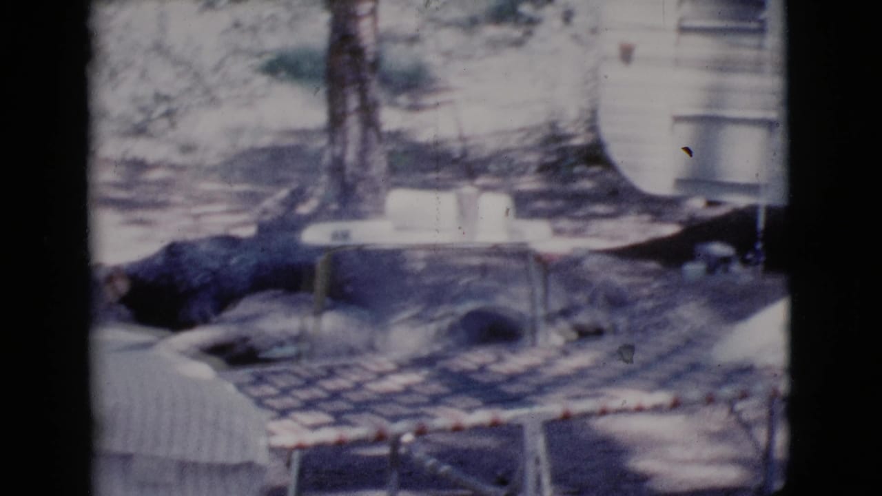 1964 Cottonwood, Arizona home-movie frame showing a white camper trailer beside a folding cot under trees.