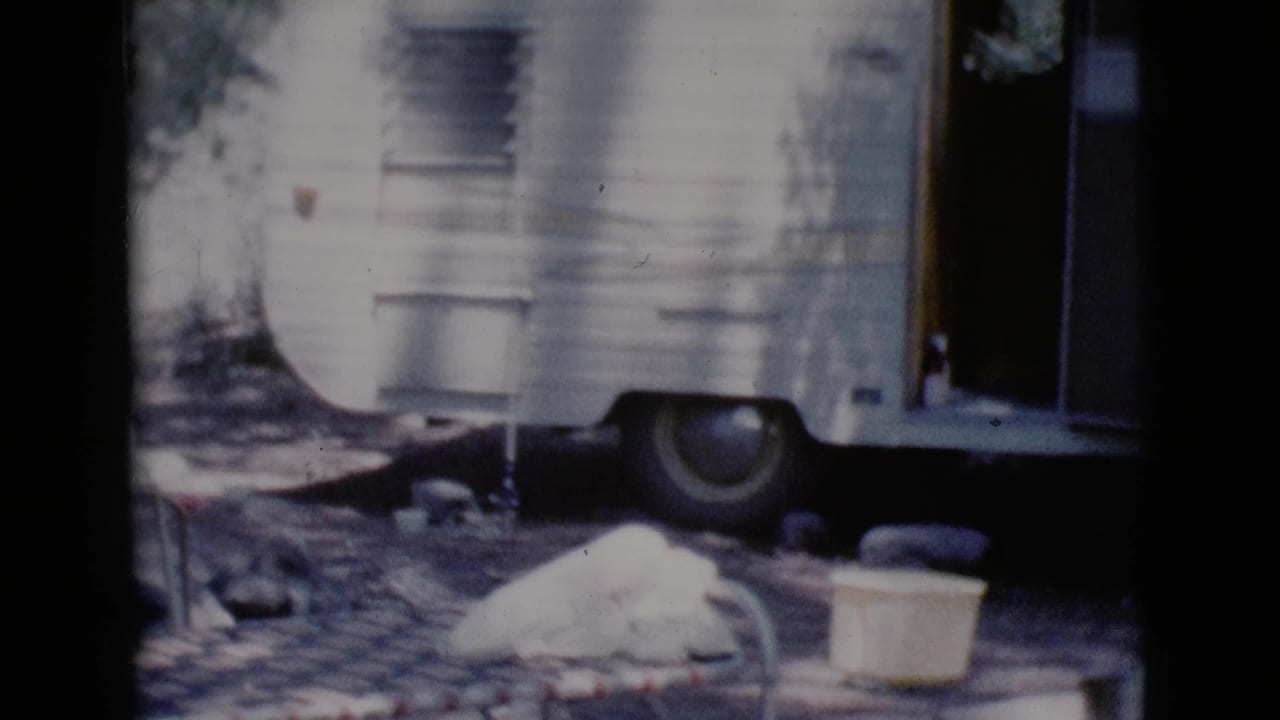 White camper trailer with open doorway, wheel, cot edge, and bucket at a campsite.