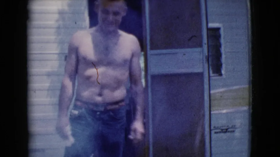 A shirtless adult man in blue jeans stands by a white camper trailer in 1964 Cottonwood, Arizona home-movie footage.