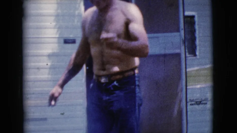 A shirtless adult man in blue jeans gestures near the camper trailer during a 1964 Cottonwood, Arizona camping scene.