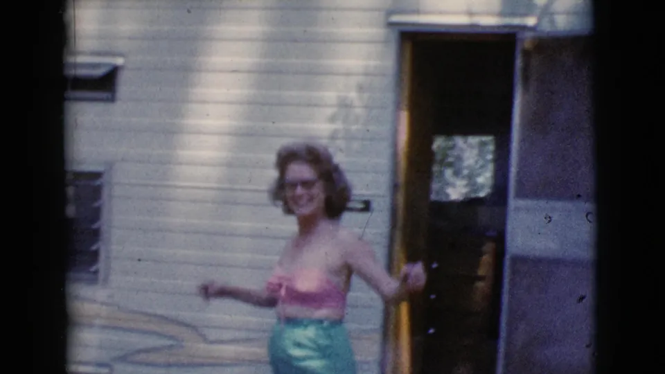 A woman steps away from the open tow camper door, smiling in dappled sunlight during the 1964 Cottonwood, Arizona home movie.