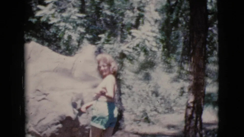 A woman in shorts stands near large rocks and trees, looking toward the camera in 1964 Cottonwood, Arizona home-movie footage.