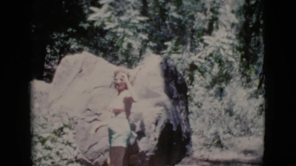The woman stands by large rocks and appears to wave or gesture toward the camera in wooded Cottonwood, Arizona footage from 1964.