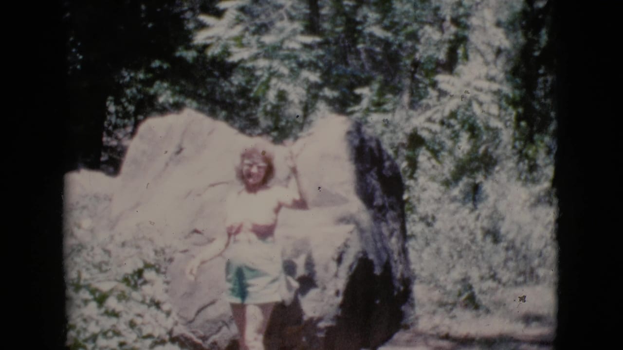1964 8mm frame from Cottonwood, Arizona, showing a woman in green shorts beside a pale boulder and trees.