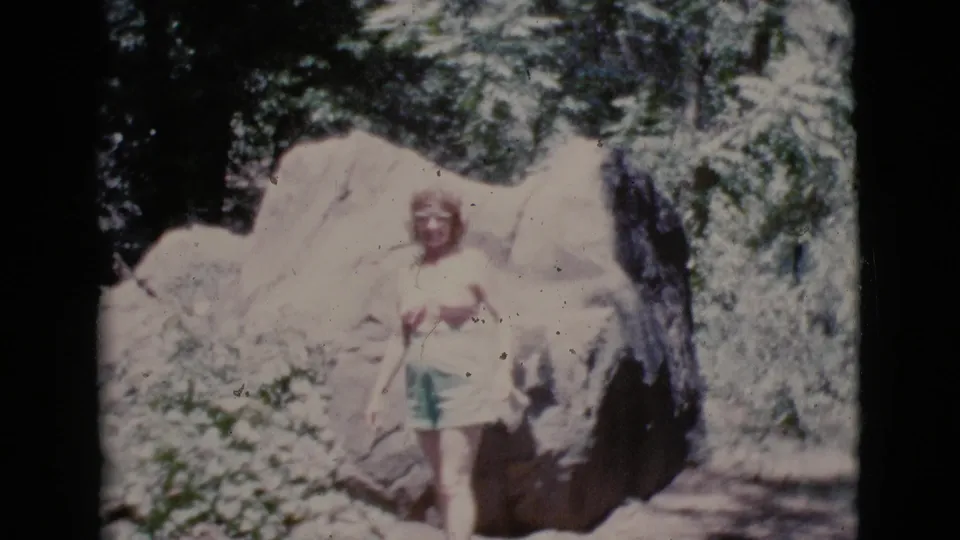 Woman in green shorts gestures toward the camera beside a large boulder in Cottonwood, Arizona, 1964 home-movie footage.