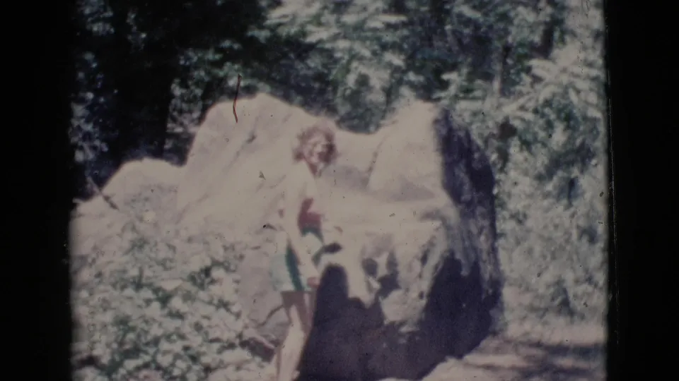 Final 1964 8mm frame with the woman beside a large boulder, trees, foliage, and a shaded path in Cottonwood, Arizona.