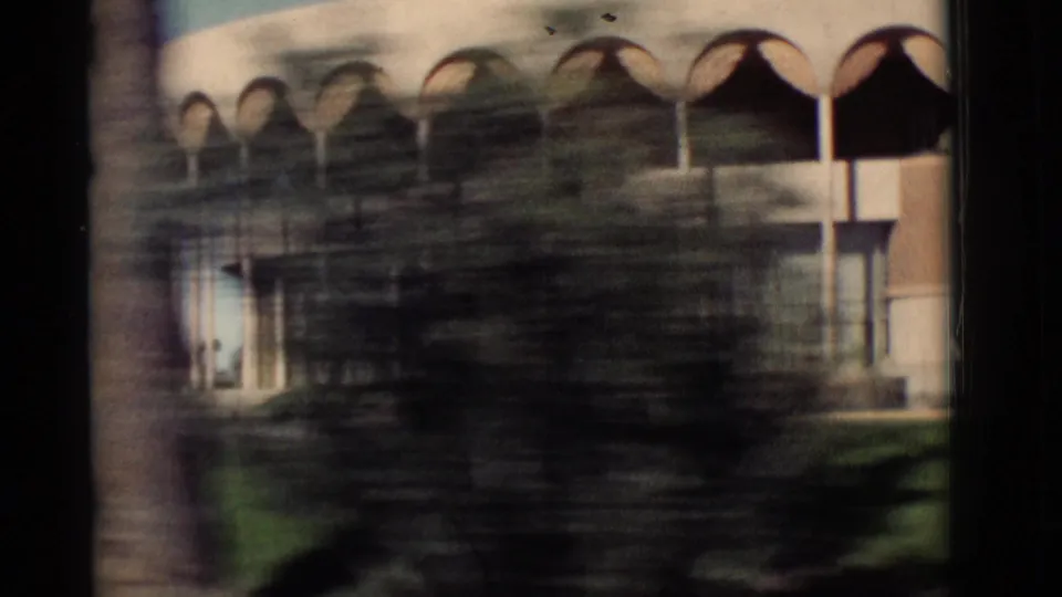 1964 New Mexico drive-by frame with the arched building partly obscured by tree blur and vehicle-window edges.