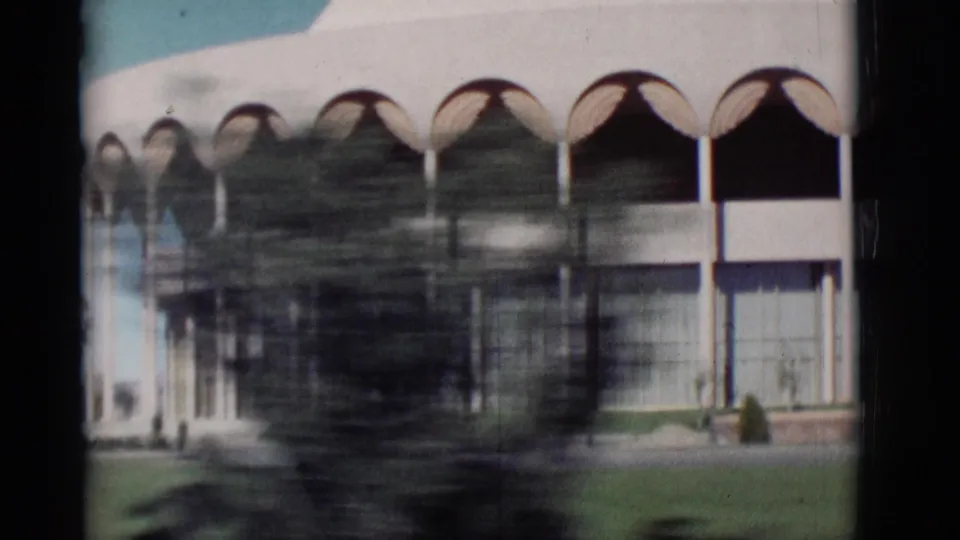 1964 New Mexico home-movie frame showing repeated arches, columns, and glass frontage interrupted by roadside tree blur.