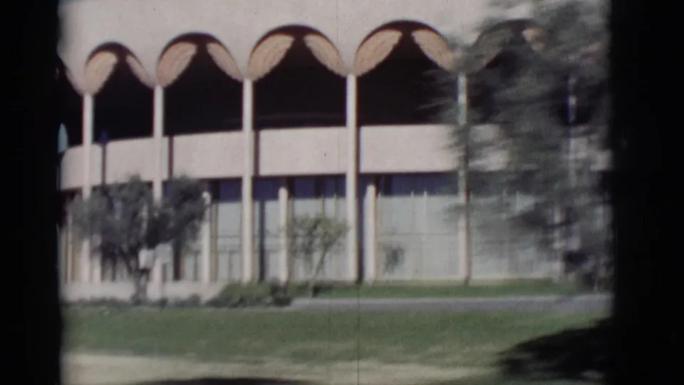 1964 New Mexico frame of a modern arched facade, broad windows, lawn, and small trees viewed from a passing vehicle.