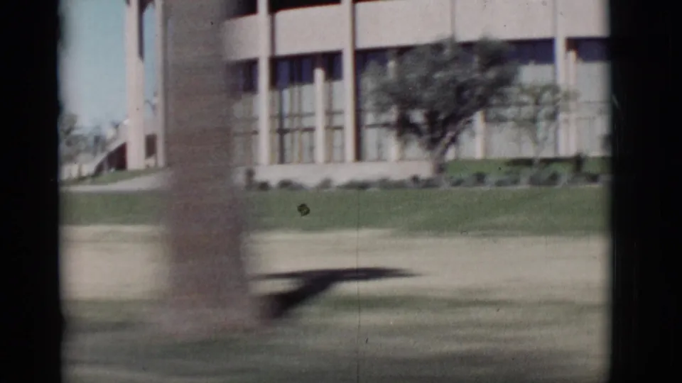 1964 New Mexico 8mm frame from a moving car, showing a modern building, green lawn, glass walls, and a blurred palm trunk.