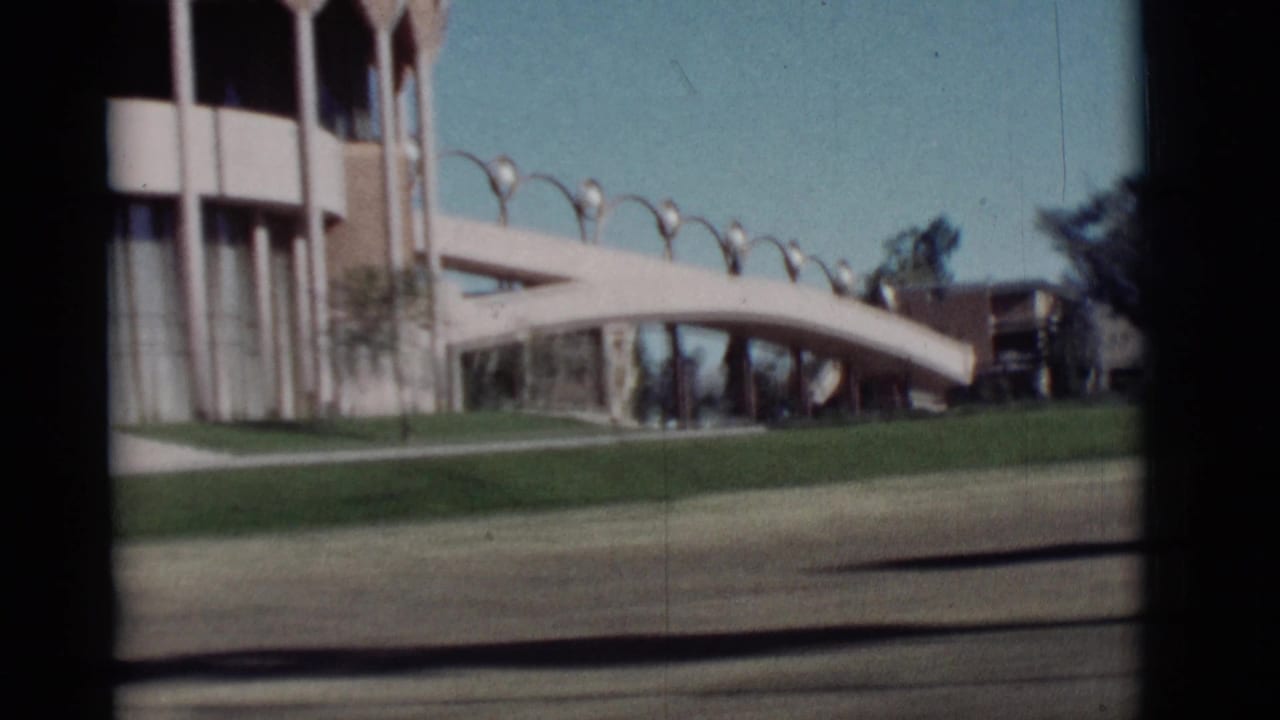 Modern building beside a curved pedestrian bridge, with lawn, columns, lamps, trees, and blue sky.