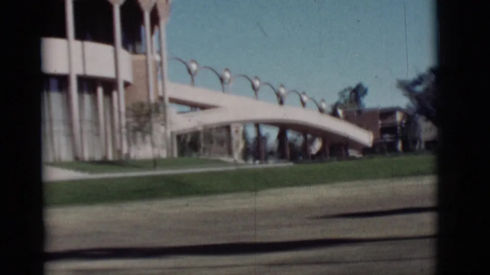 1964 New Mexico car-window frame showing a modern building with a curved pedestrian bridge, lawn, columns, and clear blue sky.