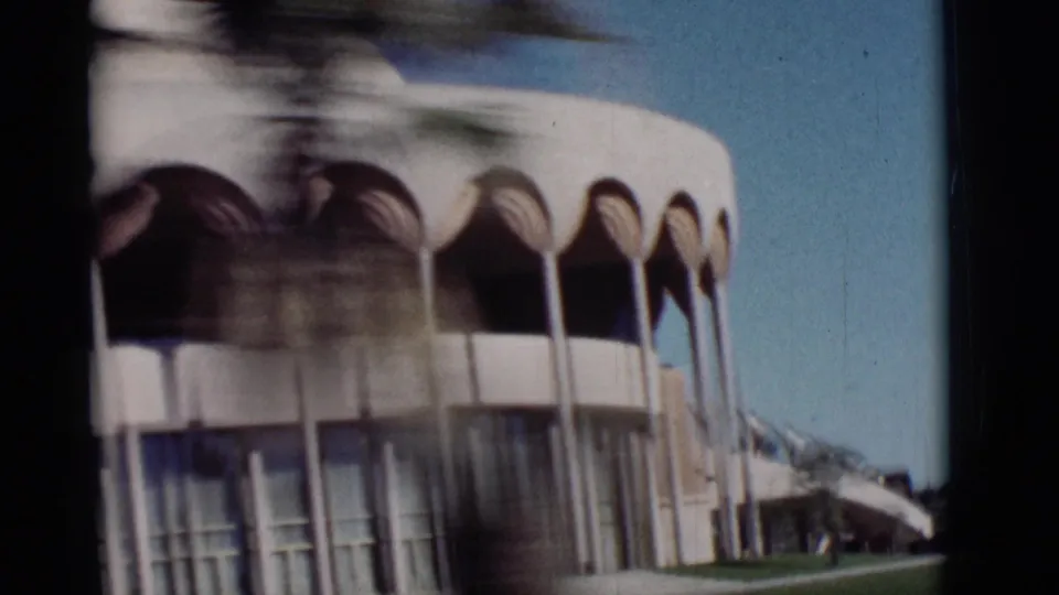 1964 New Mexico 8mm frame with motion-blurred rounded canopy forms, columns, glass frontage, lawn, and a passing tree trunk.