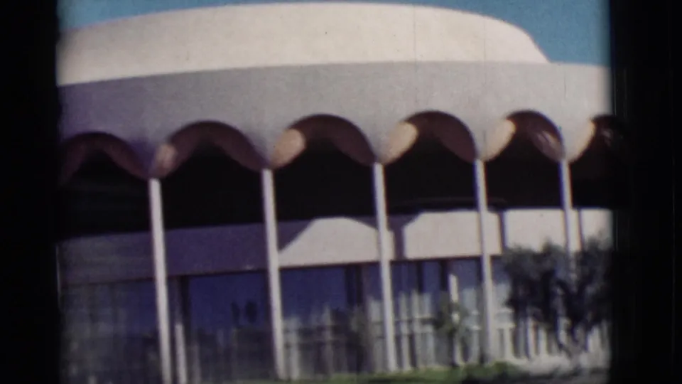 1964 New Mexico frame showing a modern facade with scalloped arches, slim columns, glass walls, palms, lawn, and blue sky.
