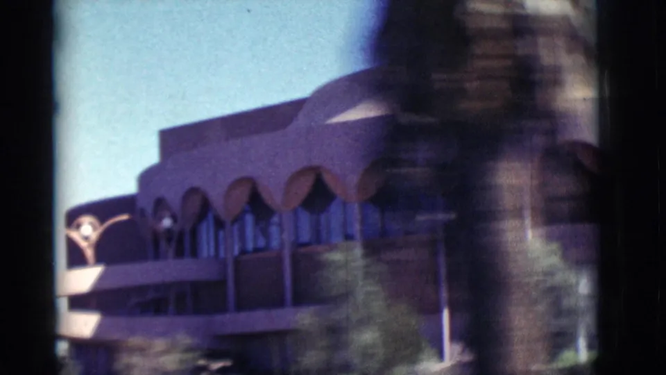 1964 New Mexico travel frame with a mid-century building, scalloped arch details, blue sky, and foreground blur from movement.