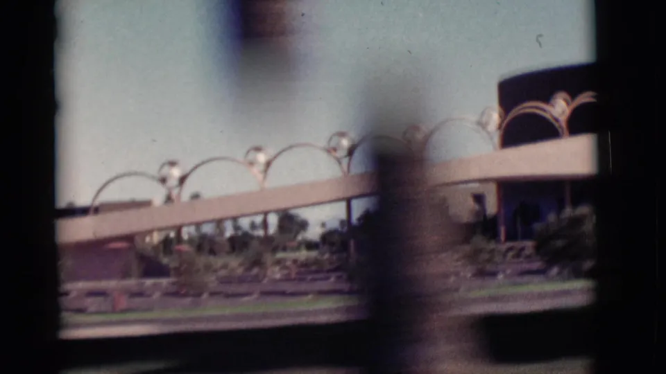 Final 1964 New Mexico travel frame showing arched architectural details, landscaped grounds, road edge, and blurred foreground from the passing car.