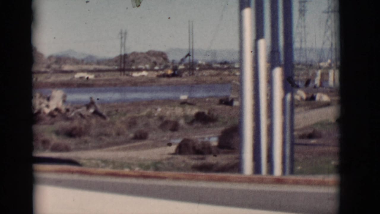 Water, scrubland, boulders, utility poles, and distant hills viewed from a moving vehicle.