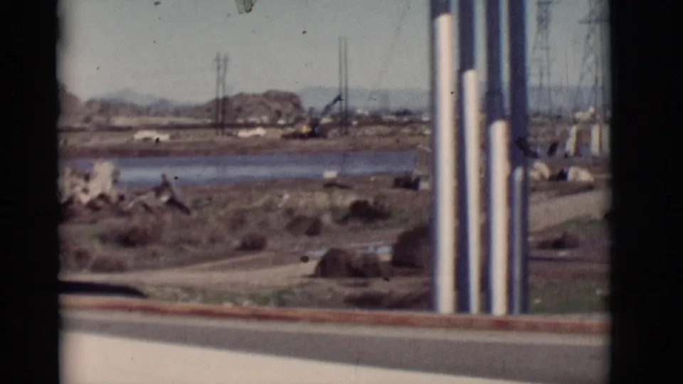 1964 New Mexico home-movie frame showing water, dry rocky terrain, utility poles, and distant hills from a roadside view.