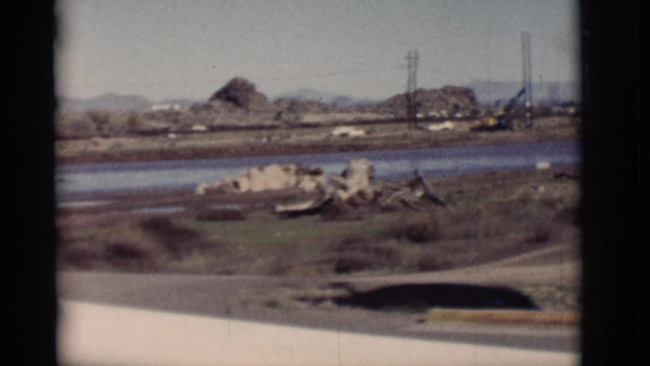 Rocky shoreline, calm water, scrub, utility poles, and low hills beyond the road edge.