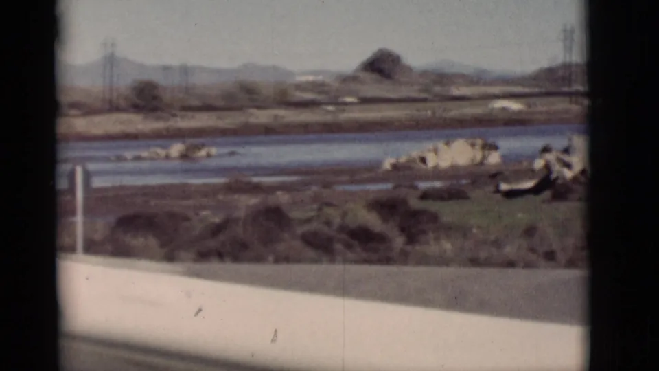 1964 New Mexico home-movie view across a waterway and dry rocky ground toward utility poles and distant hills.