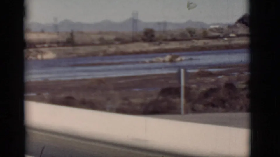 1964 New Mexico frame showing water and roadside infrastructure with scrubland, utility poles, and distant mountains.