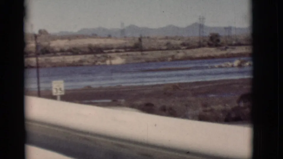 1964 New Mexico 8mm view from a bridge or vehicle window, showing a river channel, muddy flats, distant mountains, and a small sign.