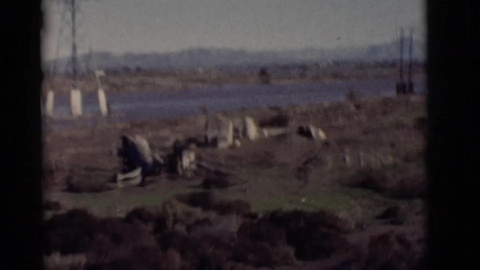 1964 New Mexico riverbank scene with transmission tower supports, scattered debris, scrubby ground, water beyond, and hazy mountains.