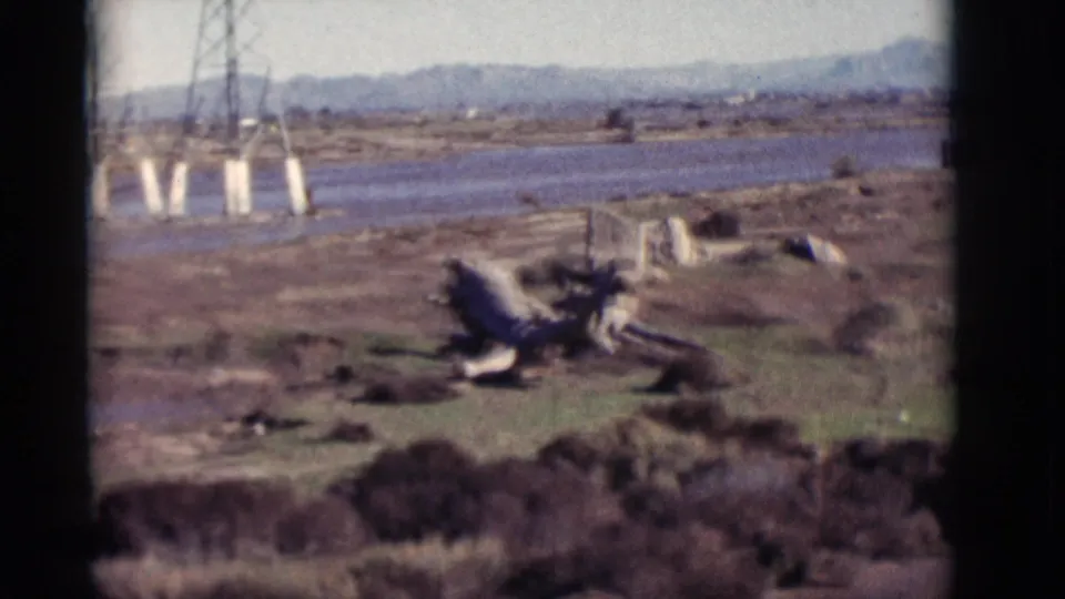 1964 New Mexico 8mm frame looking over mud flats, a river channel, utility tower supports, debris, sparse vegetation, and mountains.