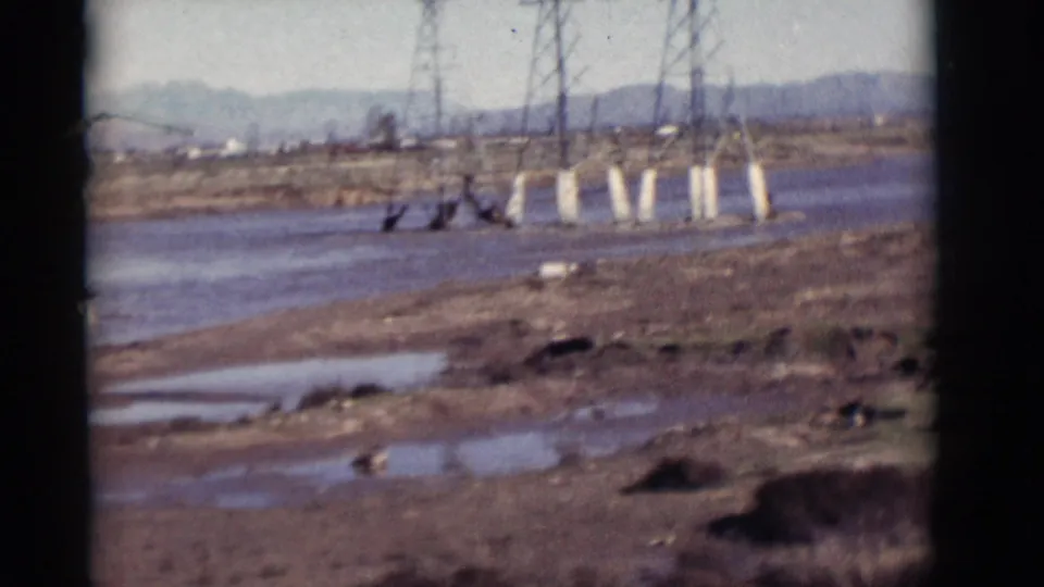 1964 New Mexico view across a shallow river and mud flats with puddles, transmission towers, scrub vegetation, and mountains in haze.