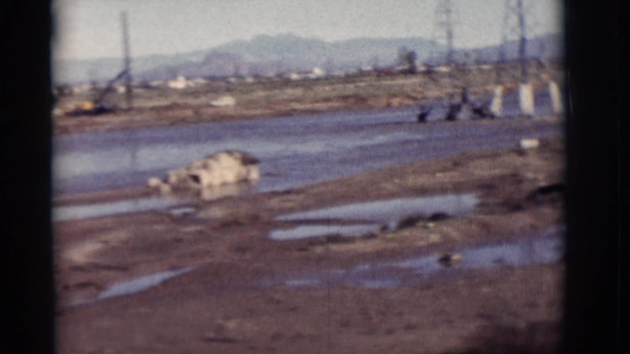 1964 New Mexico 8mm frame showing a muddy riverbank, water, scattered debris, utility towers and low mountains in the distance.