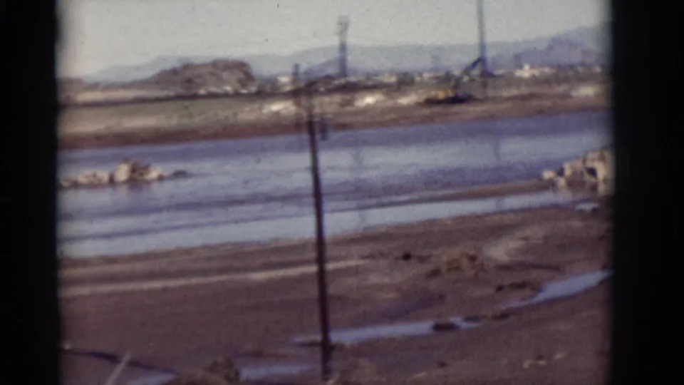 Handheld 8mm view of a New Mexico industrial riverside with water, muddy flats, utility poles, debris and low hills beyond.