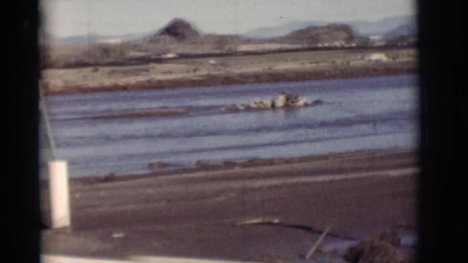 1964 New Mexico frame with water in the foreground, muddy bank, rocks or debris, utility poles, a distant mound and mountains.