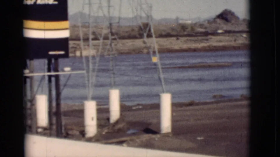 New Mexico 8mm frame showing a striped industrial structure, tower supports, white posts, water, muddy bank and distant hills.
