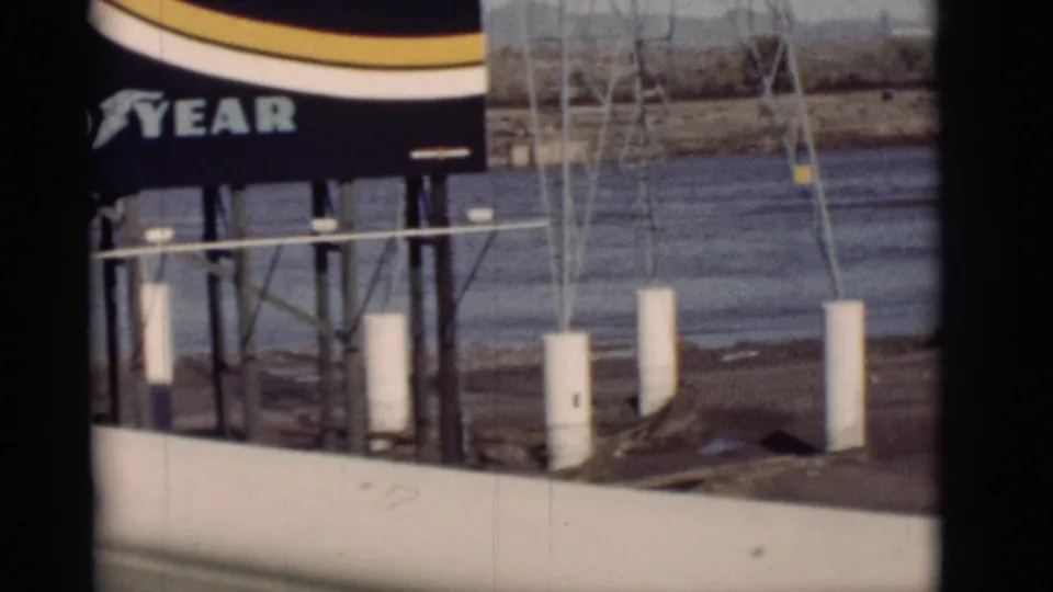 1964 New Mexico 8mm frame showing part of a Goodyear billboard beside a river, with white posts, roadside structure, and arid background.