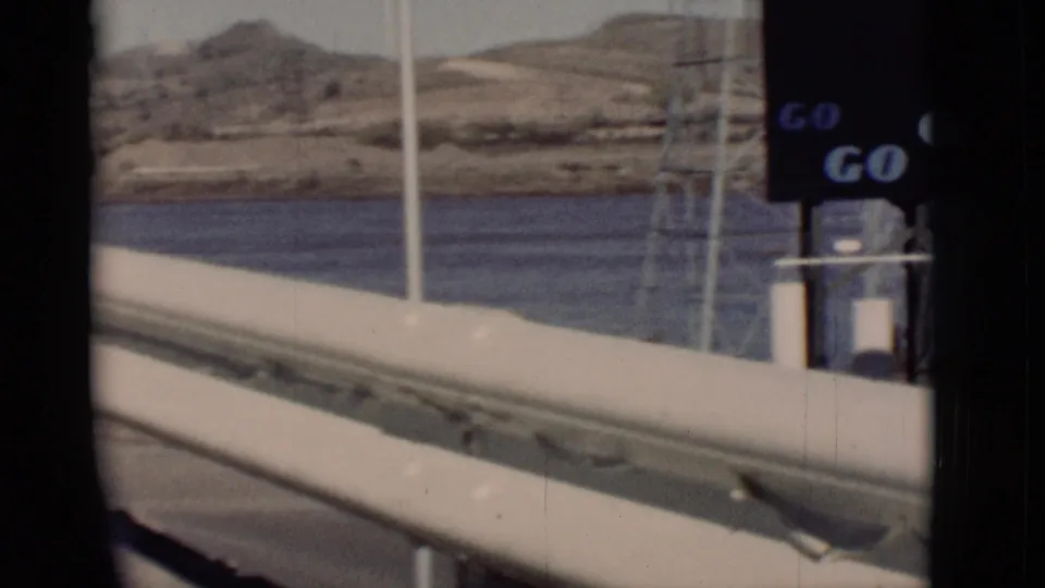 1964 New Mexico 8mm view across a railing toward river water and dry hills, with partial Goodyear sign structure at the edge.