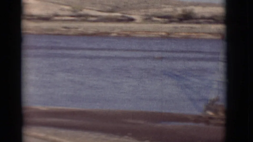 1964 New Mexico 8mm frame showing flowing river water, dry shoreline, arid hills, and a small distant person near the bank.