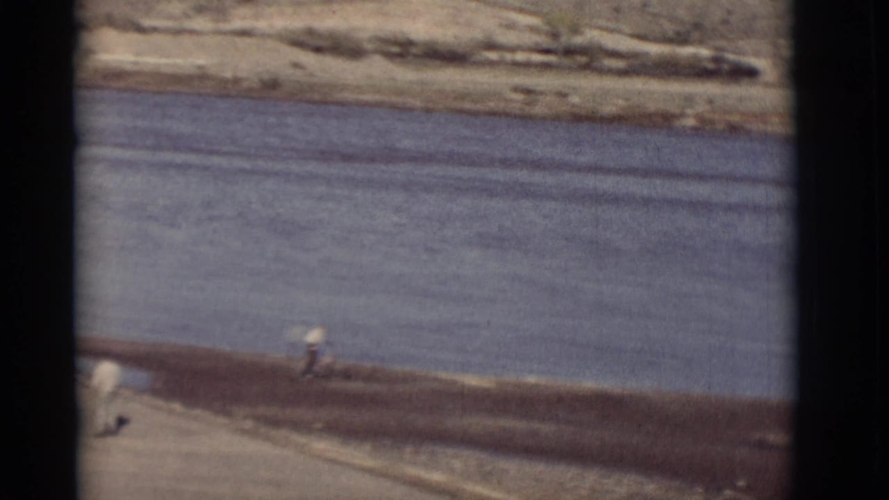 Broad river beside a dirt bank with a small figure near the water and mountains beyond.