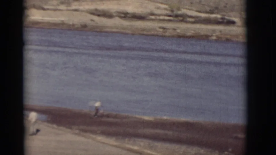 1964 New Mexico 8mm frame showing a broad river, dry roadside bank, a distant person, and pale hills beyond the water.