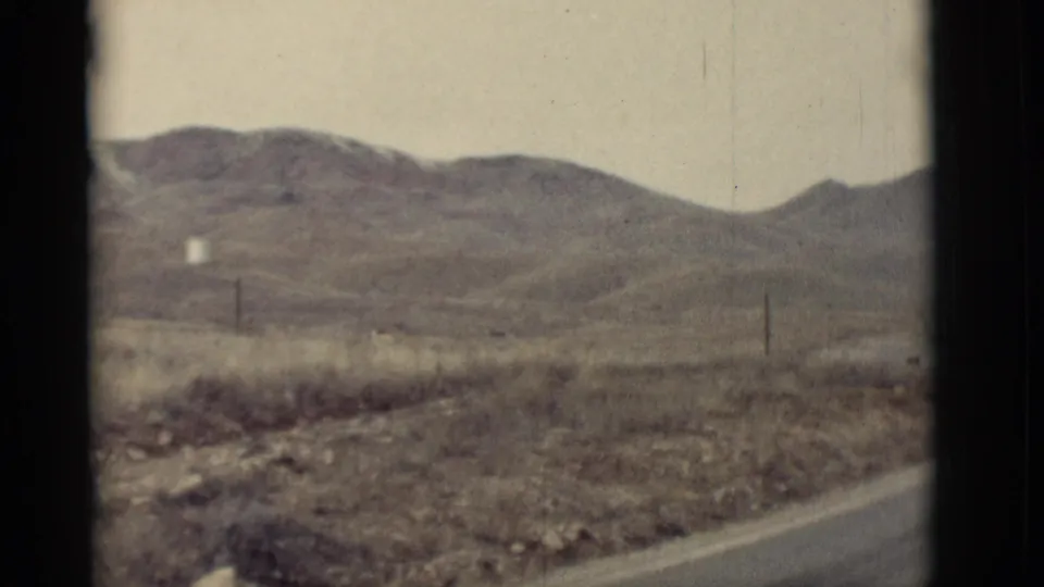 1964 New Mexico 8mm frame looking across dry roadside terrain toward low mountains and sparse fence posts.