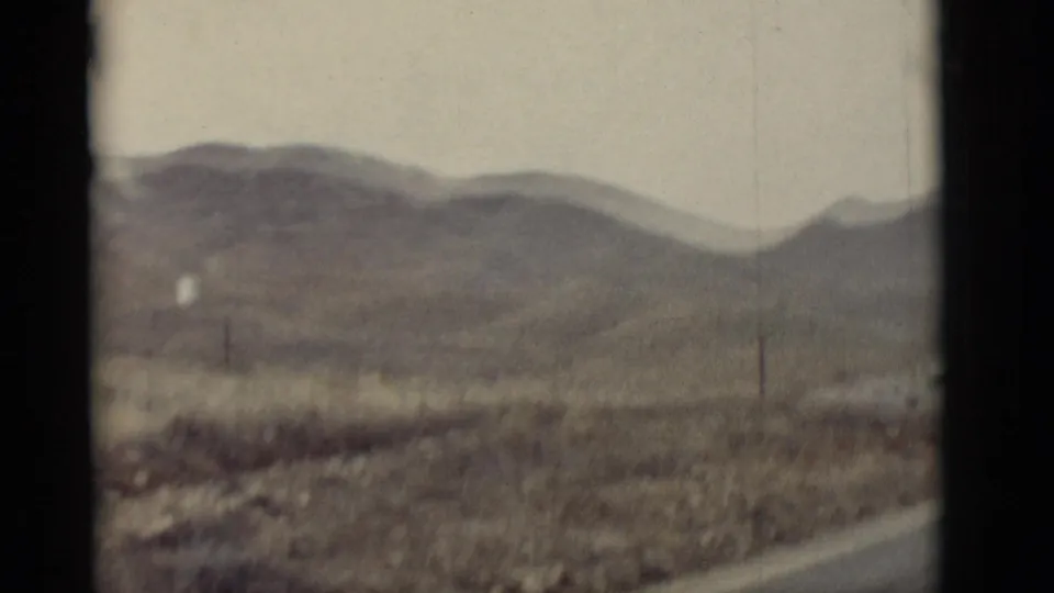 1964 New Mexico 8mm frame with soft-focus mountains, dry vegetation, fence posts, and a roadside edge.