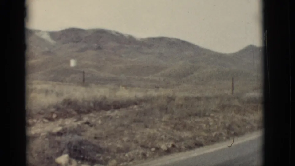 1964 New Mexico 8mm frame showing low desert hills, dry roadside ground, utility poles, and a small distant white structure.