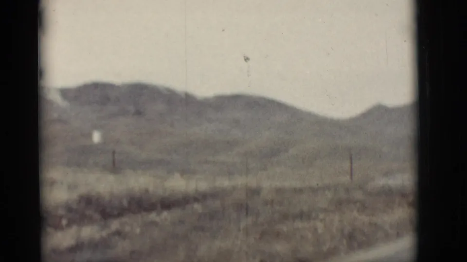 Soft 8mm view of New Mexico desert hills, dry grass, roadside ground, and a distant white structure under a pale sky.