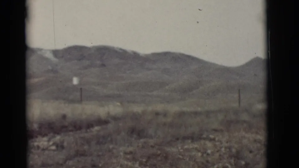 1964 8mm home-movie frame of open New Mexico desert terrain with low hills, roadside grasses, and sparse utility poles.