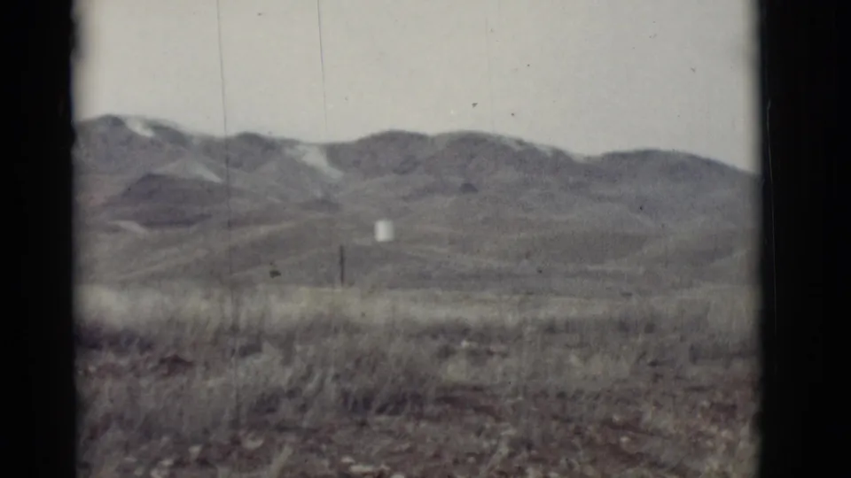 New Mexico desert landscape on 8mm film, with rolling hills, dry roadside grass, utility poles, and a small distant white structure.