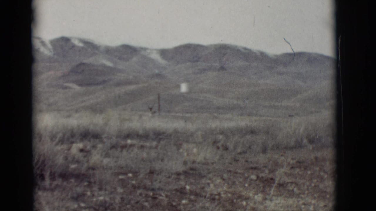 Dry grassy plain with distant low mountains and a small white object near the center.