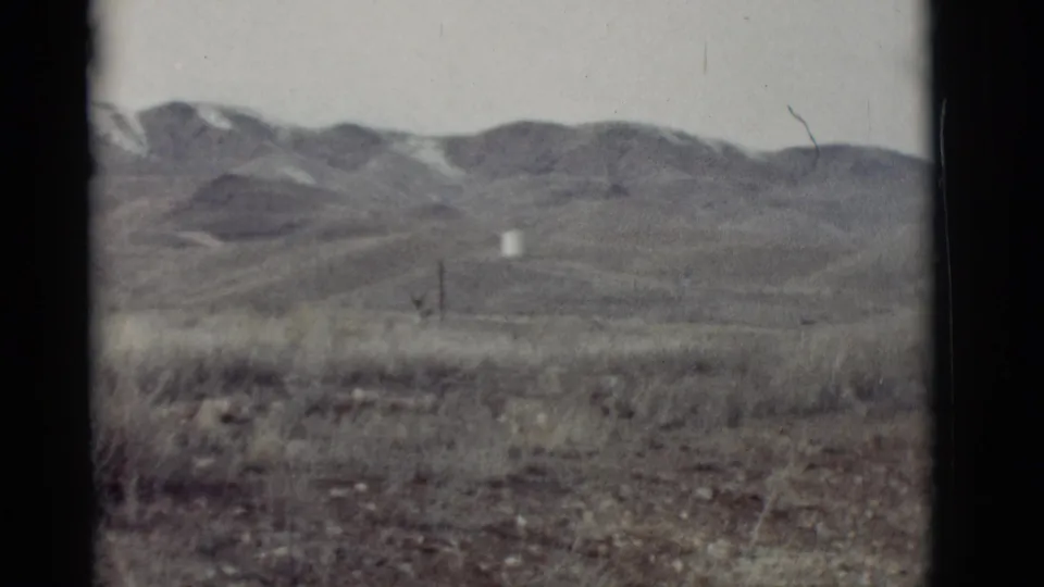1964 8mm New Mexico landscape view with dry grass, muted hills, distant snow patches, and a small white object in the middle ground.